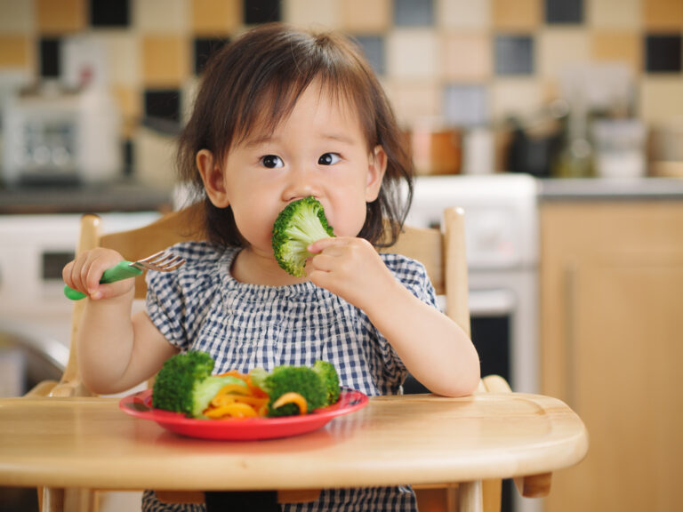 Little girl in a high chair eating carrots and broccoli at home