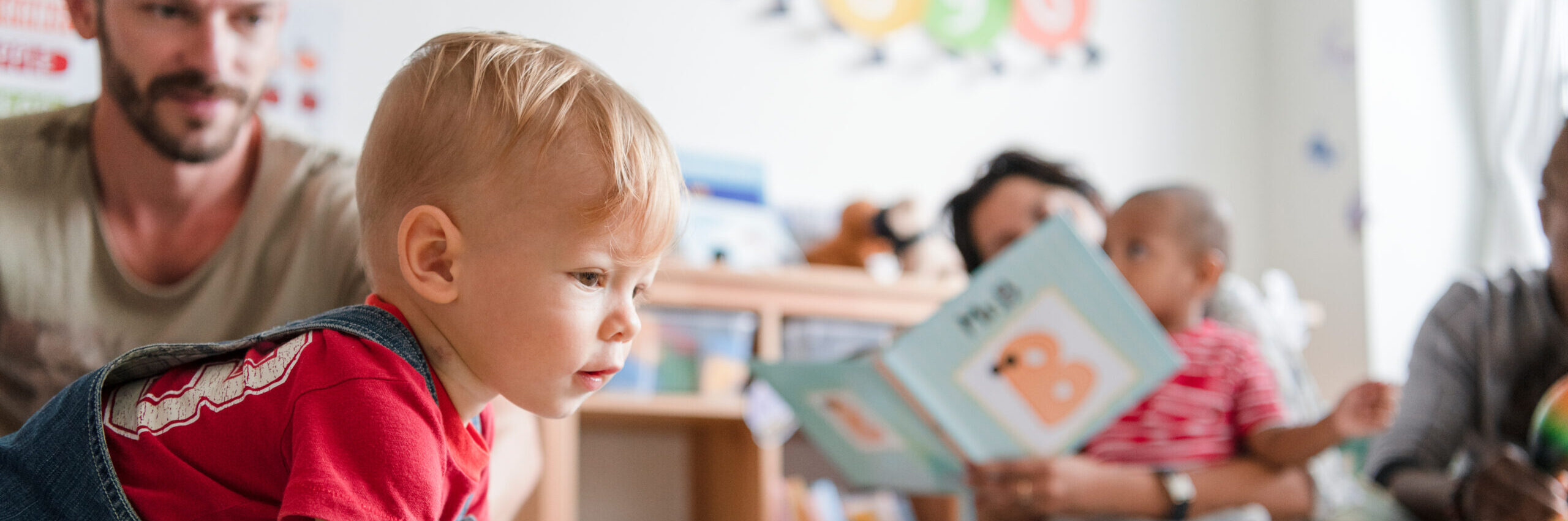 Little boy in red t-shirt crawling across the floor in a room filled with early years resources with his father in the background, and a mother reading a book with her child on her lap slightly out of focus further back
