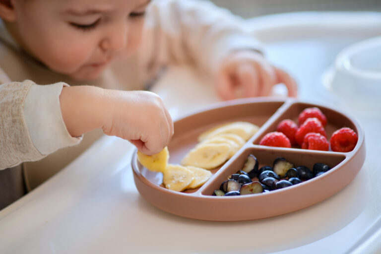 Baby at a high chair eating fruit from a sectioned wooden bowl