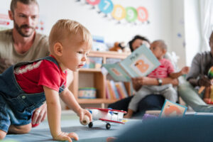 Little boy in red t-shirt crawling across the floor in a room filled with early years resources with his father in the background, and a mother reading a book with her child on her lap slightly out of focus further back