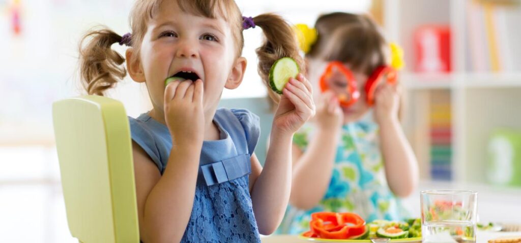 Little girl with bunches holds a cucumberin her mouth and another cucumber in her hand
