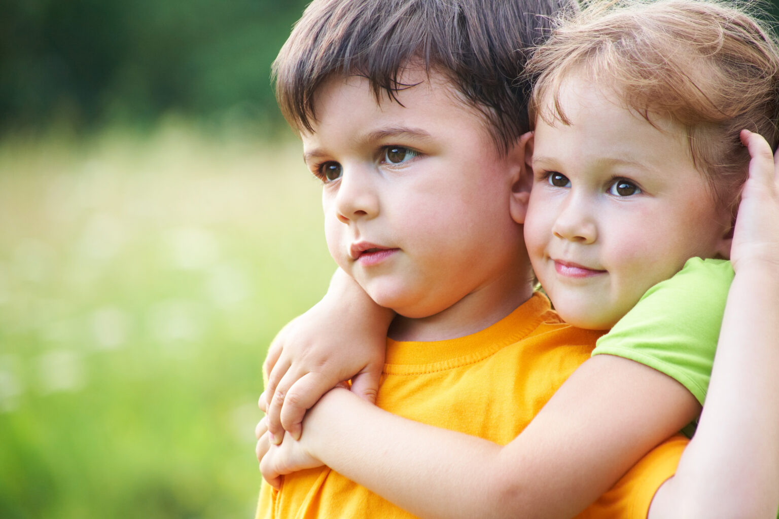 Little boy staring into the distance in a field with a younger girl hugging him from behind