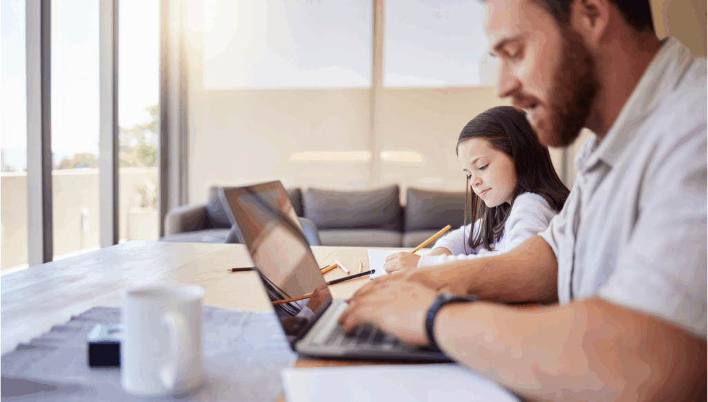 Adult man on a laptop sat next to a young girl writing at a table