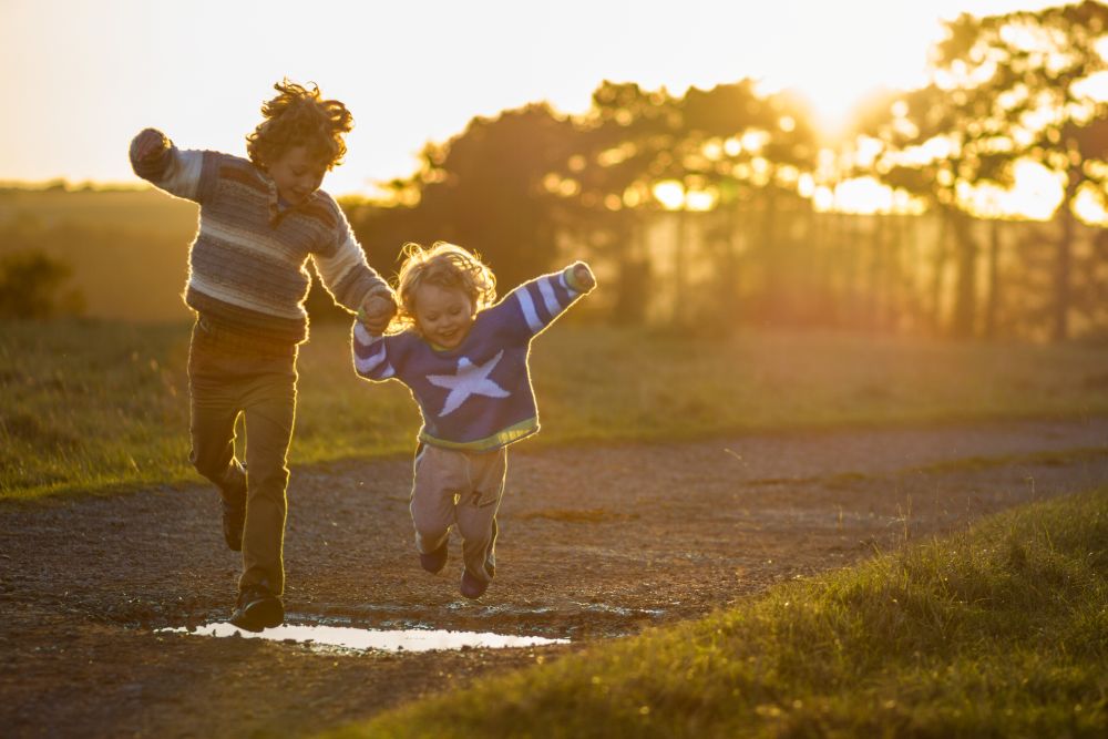 Little boy in a blue jumper holding hands with his older brother and jumping over a puddle on a path in the middle of two strips of grass as the sun sets in the background