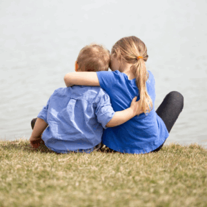 Girl sat next to boy hugging around his shoulders looking out into the sea
