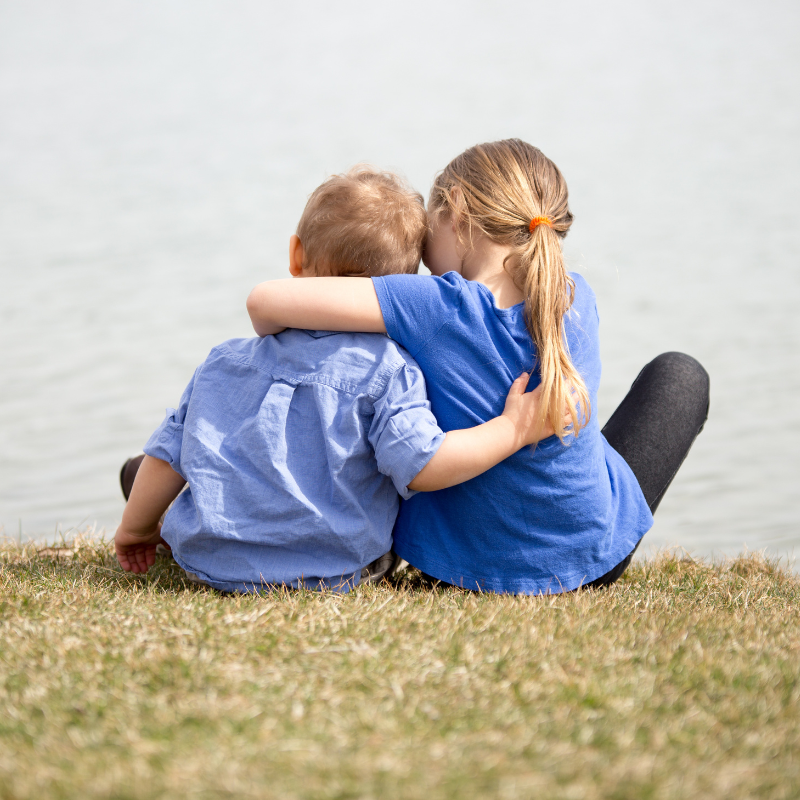 Girl sat next to boy hugging around his shoulders looking out into the sea