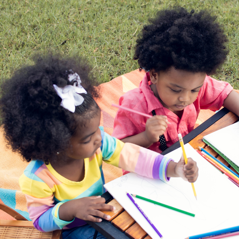 Little boy and girl sat on the grass writing with coloured pens on paper