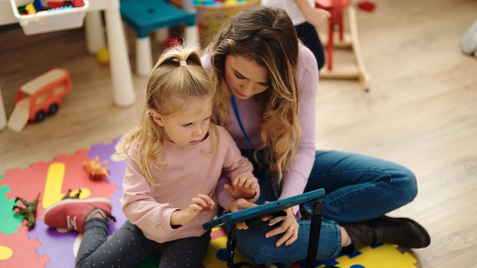 Educator sat with girl on the floor of an early years setting, sharing a digital tablet