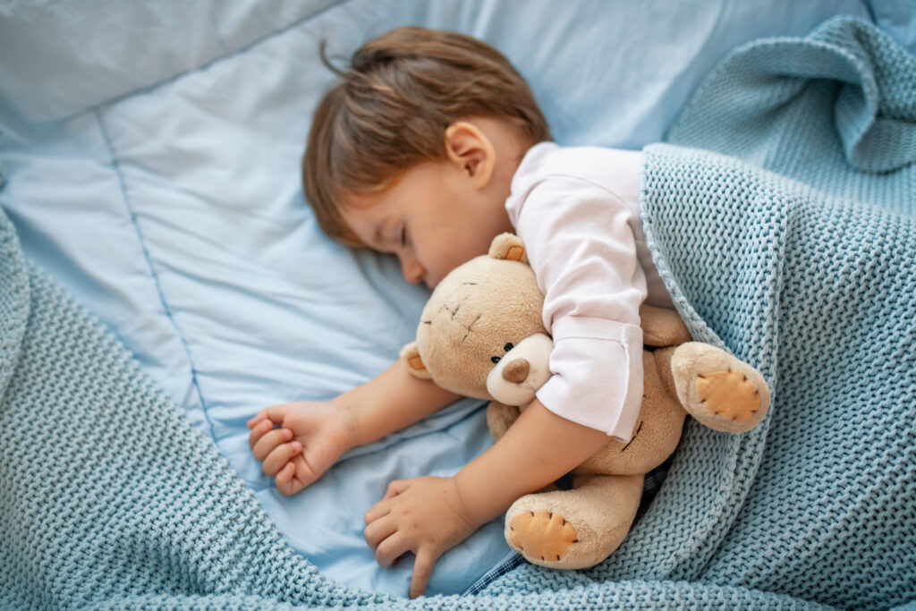 Little boy sleeping on his side cuddling a teddy bear under a blue blanket