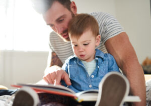 Toddler in a blue shirt sat on his dad's lap reading a book