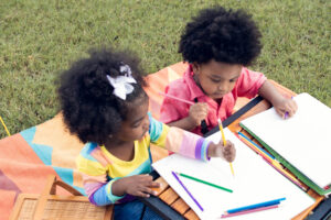 Two young children sat outside writing on paper with a range of coloured pencils
