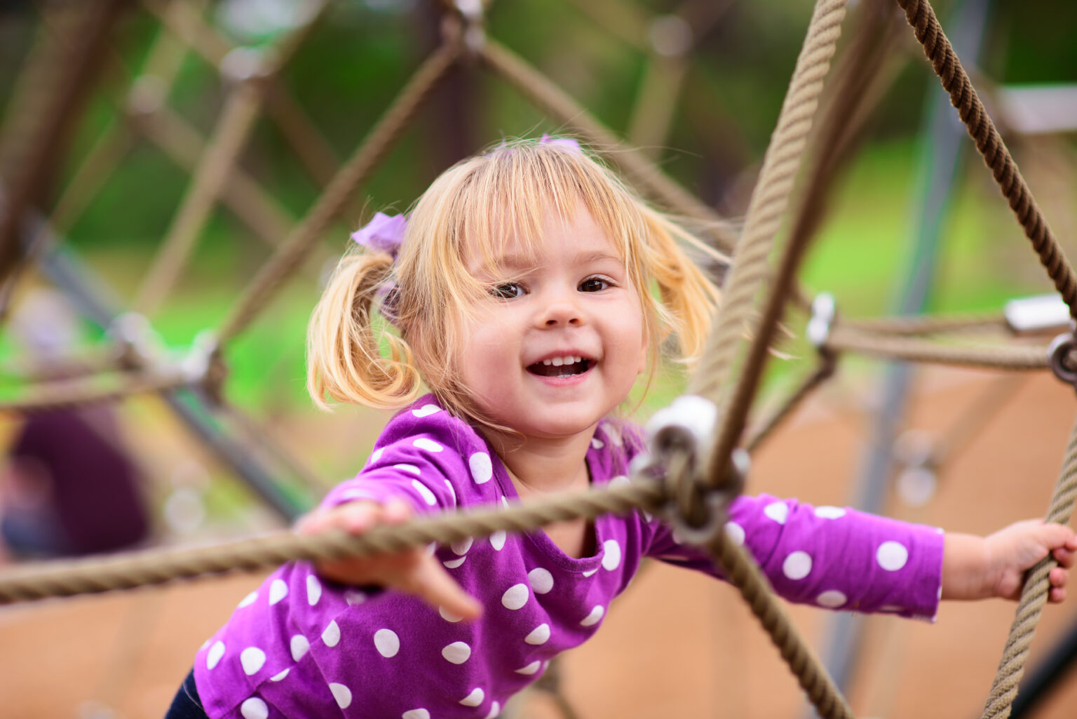 Little girl with pigtails smiling, holding onto ropes in a climbing frame
