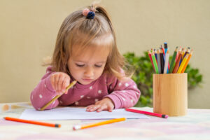Little girl in a pink top with white spots sat a table with pencils and paper in from of her considering what to draw