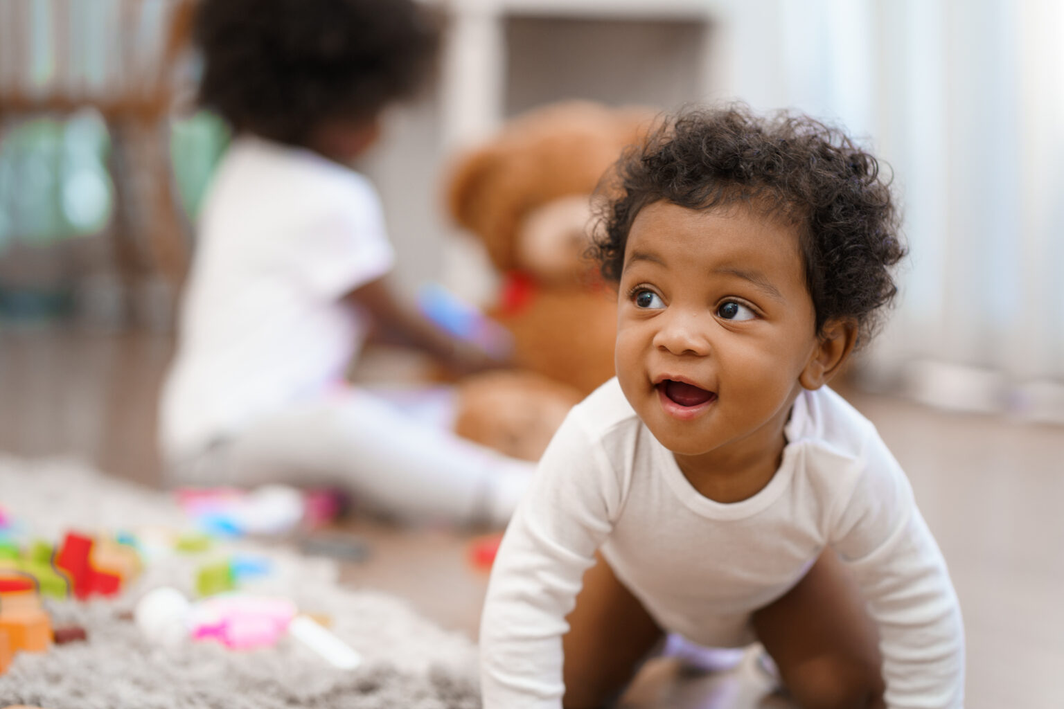 Smiling little boy crawling with another baby in the background out of focus playing with a teddy