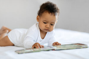 Baby lying on front on bed with his fingers on the pages of a board book