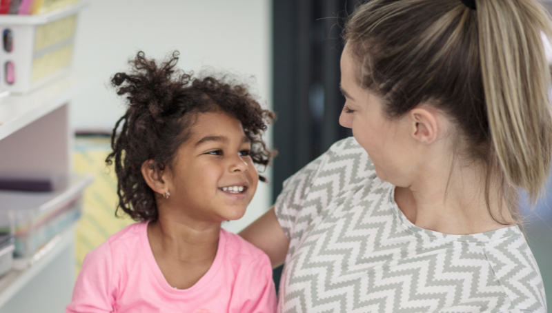 Little girl in a pink top sat on an adult woman's lap smiling up at her