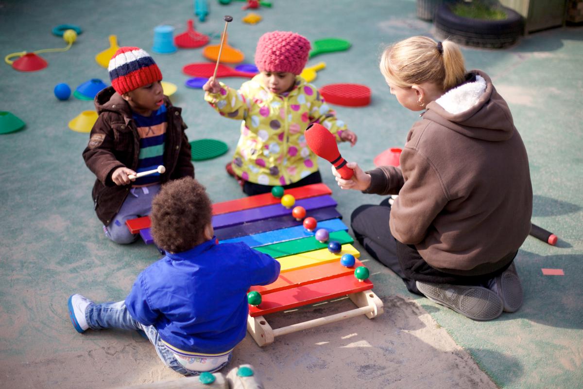 Three children play with a xylophone outdoors, whilst an adult watches