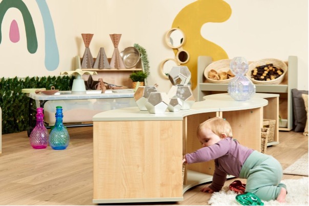 toddler in green trousers and purple top explores underneath a play bench in what looks like a baby room