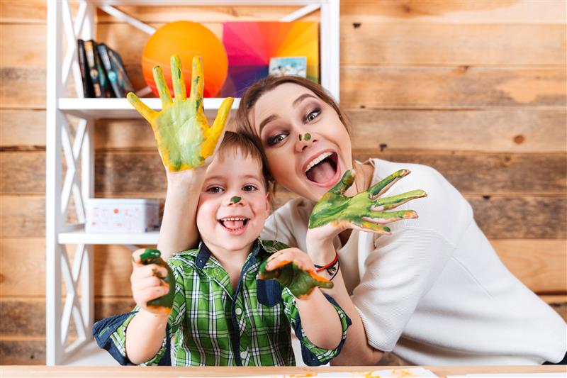 A woman and a boy sat by a table with green and yellow paint on their hands and noses. They are both smiling wide. The woman is wearing a while long sleeved top and the boy is wearing a green tartan button-up shirt.