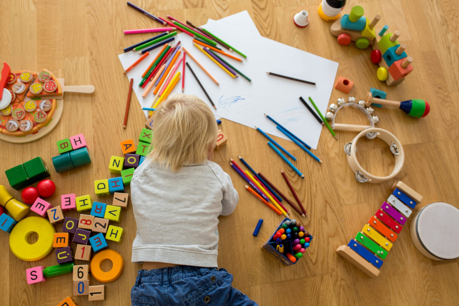 Little toddler boy lying on the floor drawing with pastels and coloring pens, playing with early development wooden toys