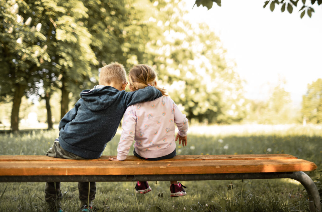 Little boy and girl sat on a bench facing away from the camera, with the boy's arm around the girls shoulder