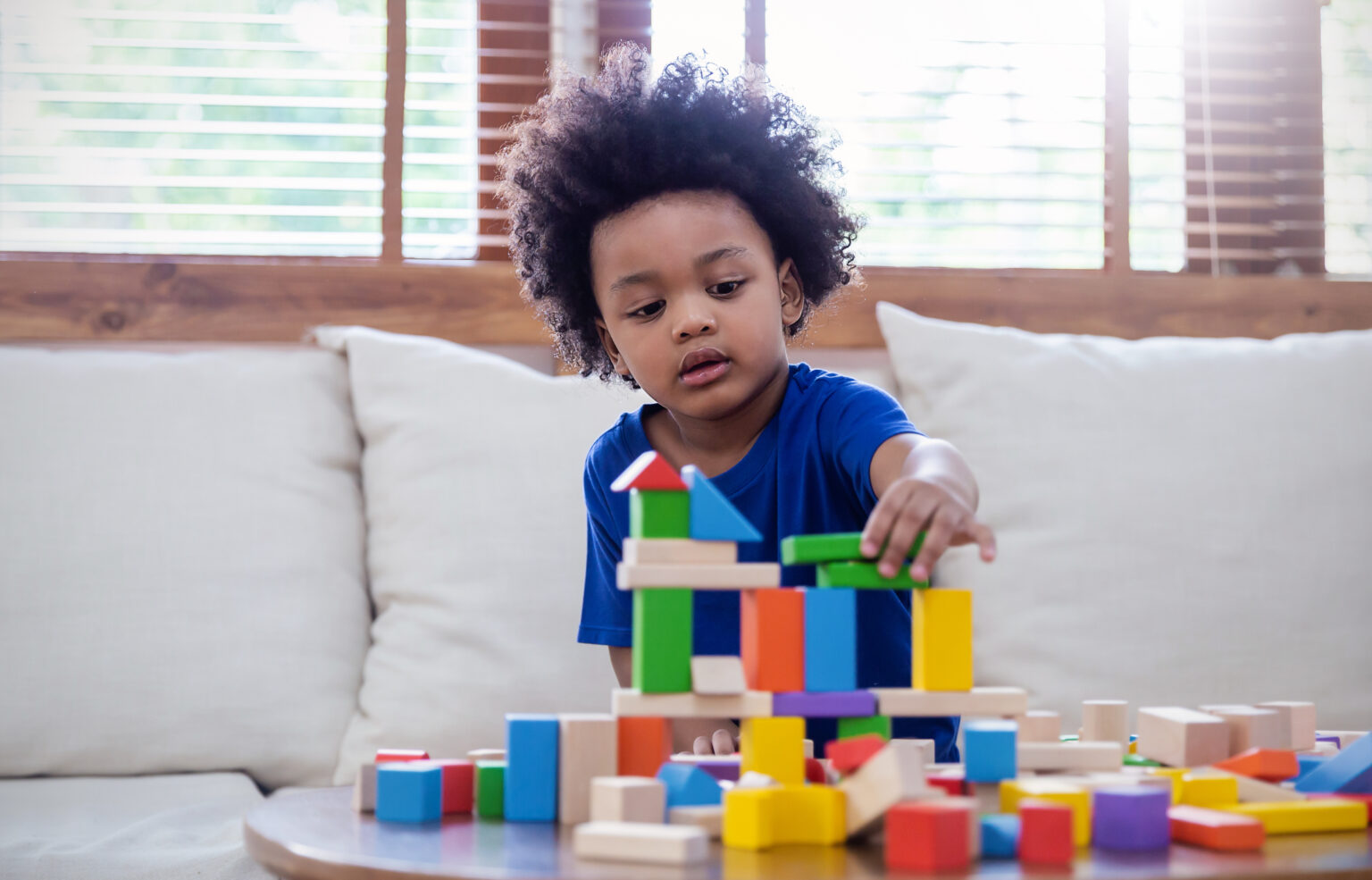 Little boy on cream sofa carefully building a structure out of colourful wooden blocks on the coffee table in front of him
