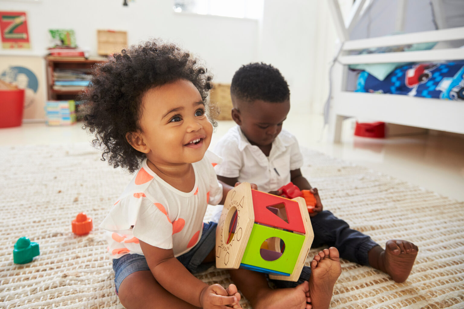 Llittle girl sat smiling on a carpet with a wooden shape sorter in her hands, next to a little boy