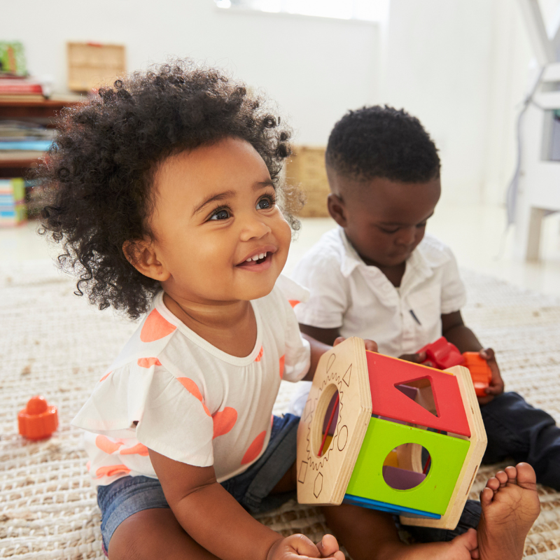 Llittle girl sat smiling on a carpet with a wooden shape sorter in her hands, next to a little boy