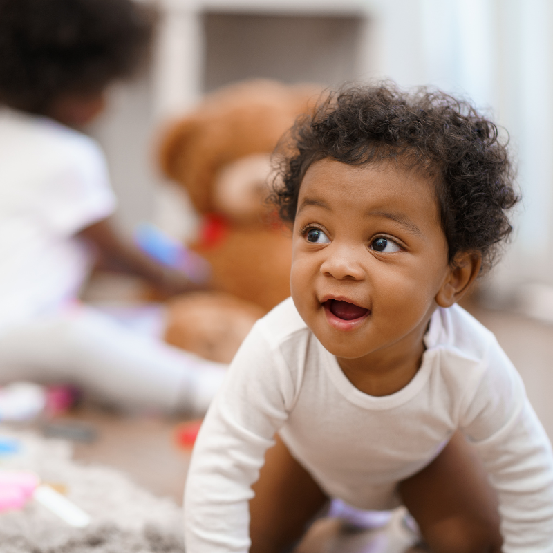 Smiling little boy crawling with another baby in the background out of focus playing with a teddy