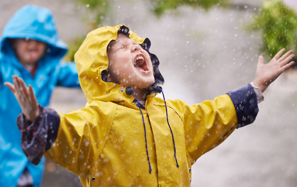Little boy in a yellow rain mac playing in the rain, with another boy in a blue rain mac playing behind him