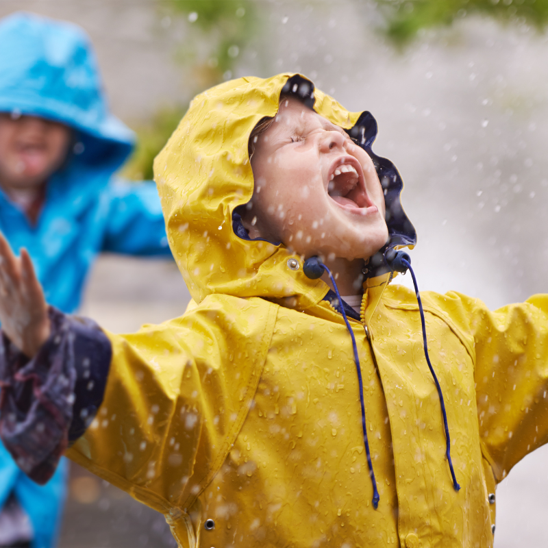 Little boy in a yellow rain mac playing in the rain, with another boy in a blue rain mac playing behind him