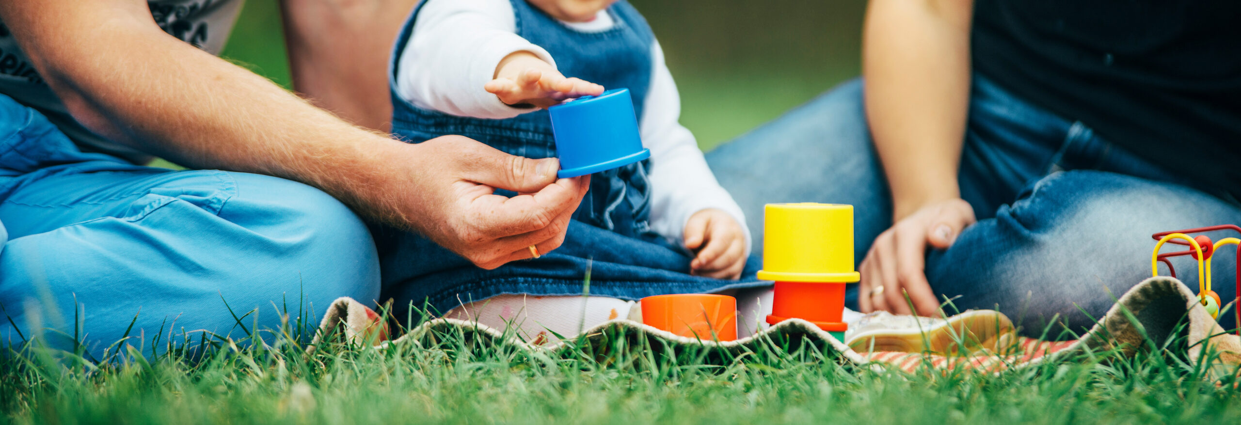 Toddler sat on the grass with her parents stacking colourful cups