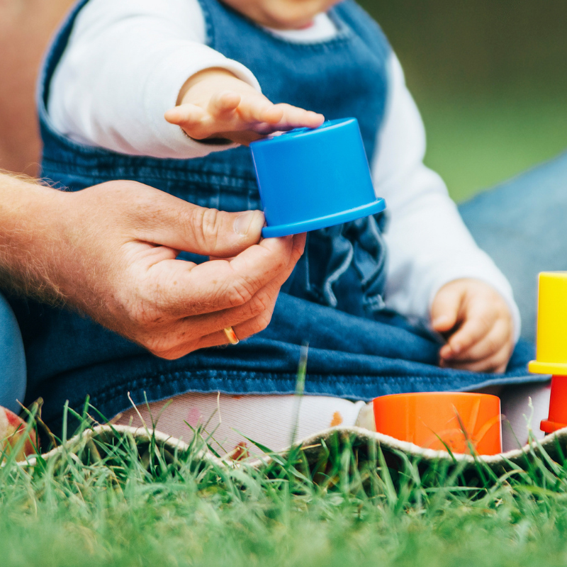 Toddler sat on the grass with her parents stacking colourful cups
