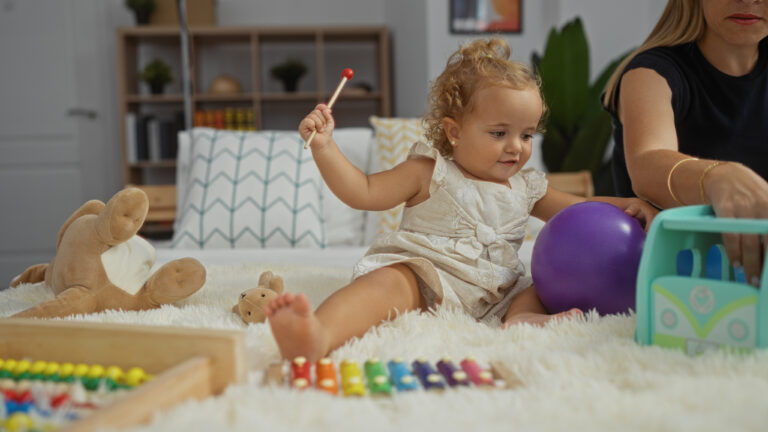 Little girl with blonder curly hair plays with xylophone on fluffy white rug