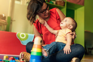 Baby in yellow t-shirt sits on mother's knee, looking at each other with smiles, with a stack of colourful blocks in front of them