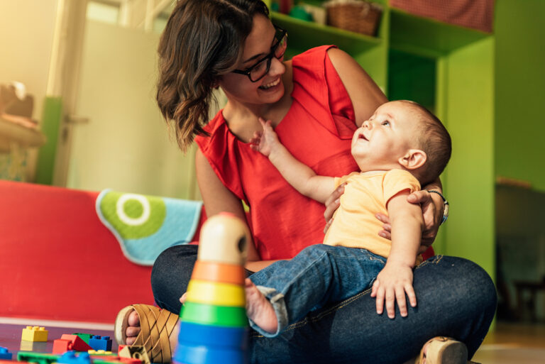 Baby in yellow t-shirt sits on mother's knee, looking at each other with smiles, with a stack of colourful blocks in front of them