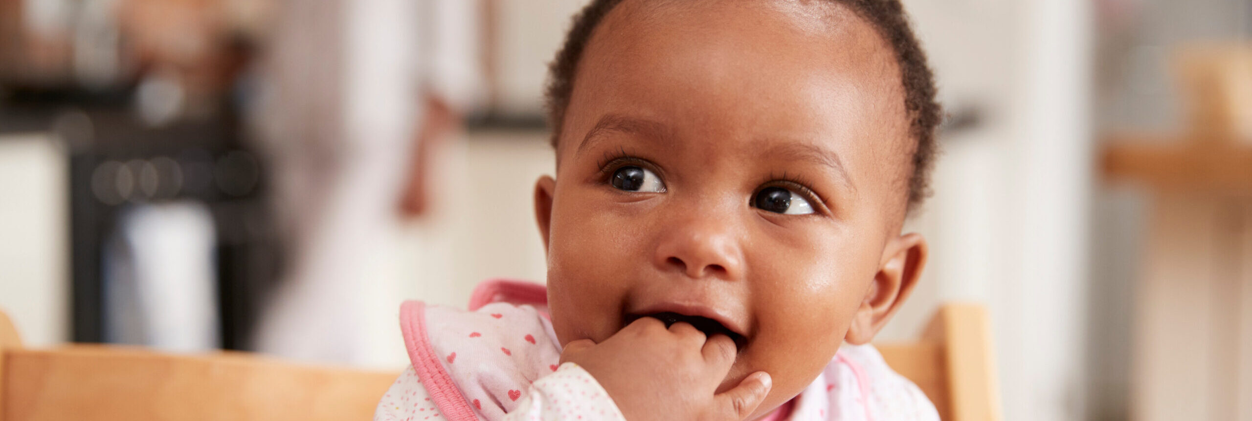 Cute Baby Girl Wearing Bib Sitting In High Chair Close up of a smiling baby with her fingers in her mouth and wearing a pink bib sat in a wooden high chair