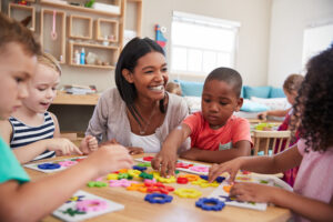 Nursery teacher sits smiling with a group of four young children as they make pictures with colourful shapes