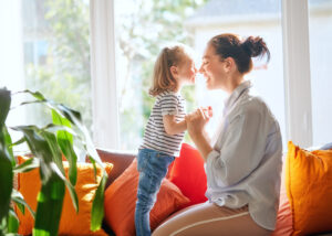 Little girl in stripy tshirt and mother hold hands and touch noses together in front of sunny window