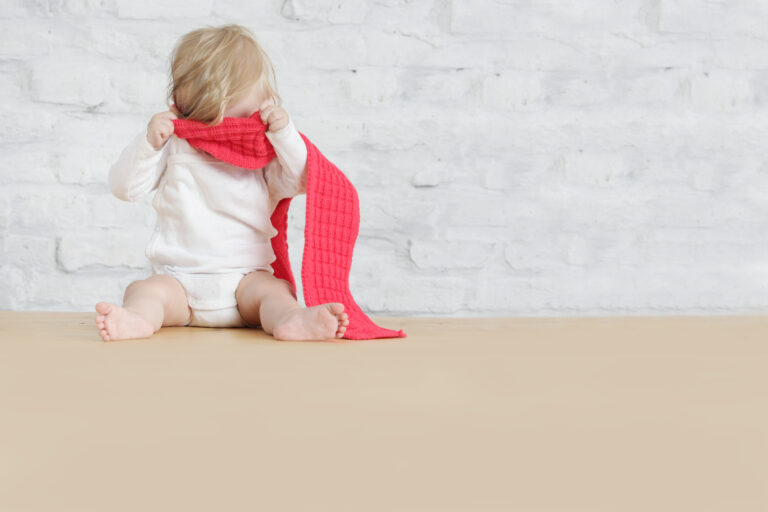 Toddler wearing white long-sleeved romper and sat in empty room with pinky-red scarf wrapped round eyes