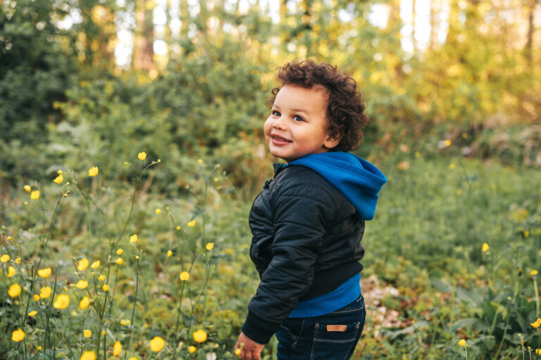 Young mixed race boy stood in grassy area beside woodland, wearing a black jacket over a blue hoodie half-turning round to the left and smiling.