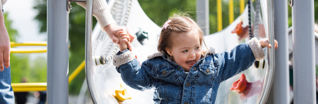 Little girl in a demin jacket and jeans smiling coming to the bottom of a metal slide in a playground holding the hand of an adult woman who is mostly out of shot