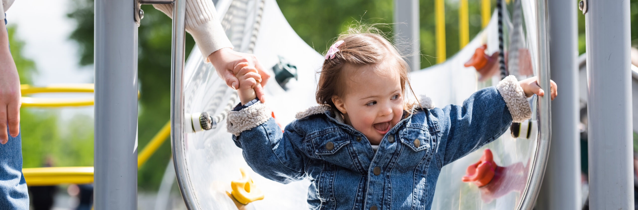 Little girl in a demin jacket and jeans smiling coming to the bottom of a metal slide in a playground holding the hand of an adult woman who is mostly out of shot