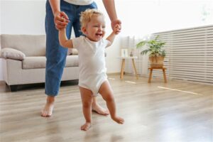 Smiling toddler in a white onesie being supported to walk across their living room floorboards by an adult whose top half is out of shot