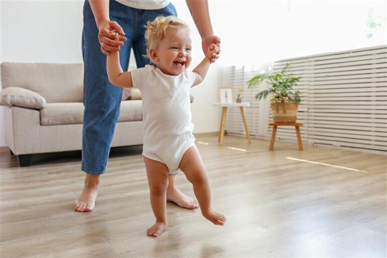 Smiling toddler in a white onesie being supported to walk across their living room floorboards by an adult whose top half is out of shot
