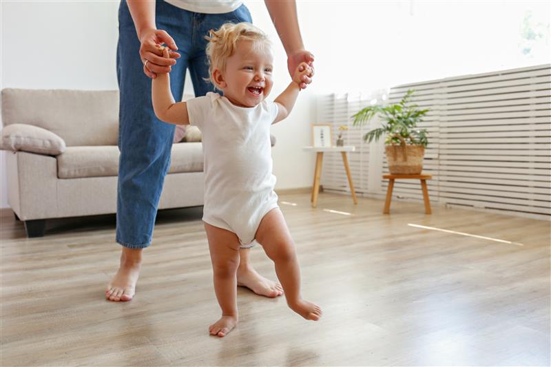 Smiling toddler in a white onesie being supported to walk across their living room floorboards by an adult whose top half is out of shot