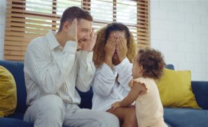 A mother and father sat on a blue sofa playing peekaboo with a young toddler girl leans forward with her hands on the mother's knees