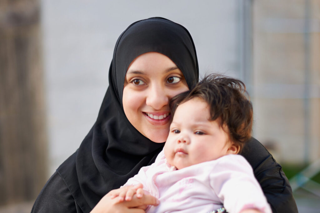 Mother wearing a black hijab smiling holding her baby daughter in front of her