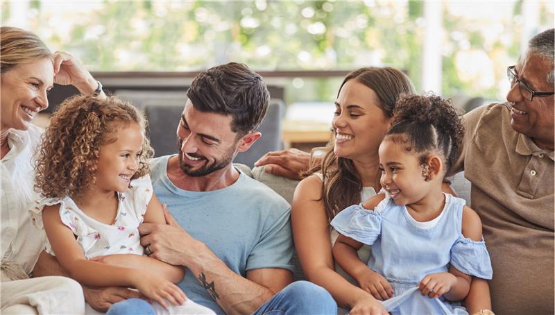 A family sit on a sofa and smile at eachother. A woman sits on the end, followed by a young girl, a man, a woman with a young girl on her lap and another man next to them