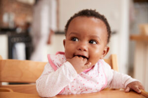 Close up of a smiling baby with her fingers in her mouth and wearing a pink bib sat in a wooden high chair